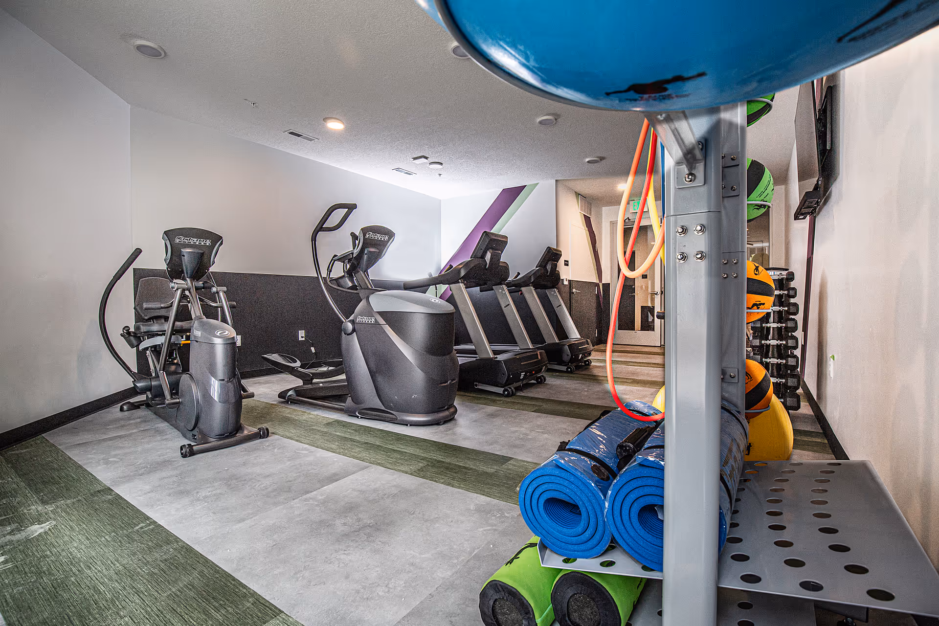 Small fitness room with treadmills and elliptical machines and a rack of yoga mats and exercise balls.