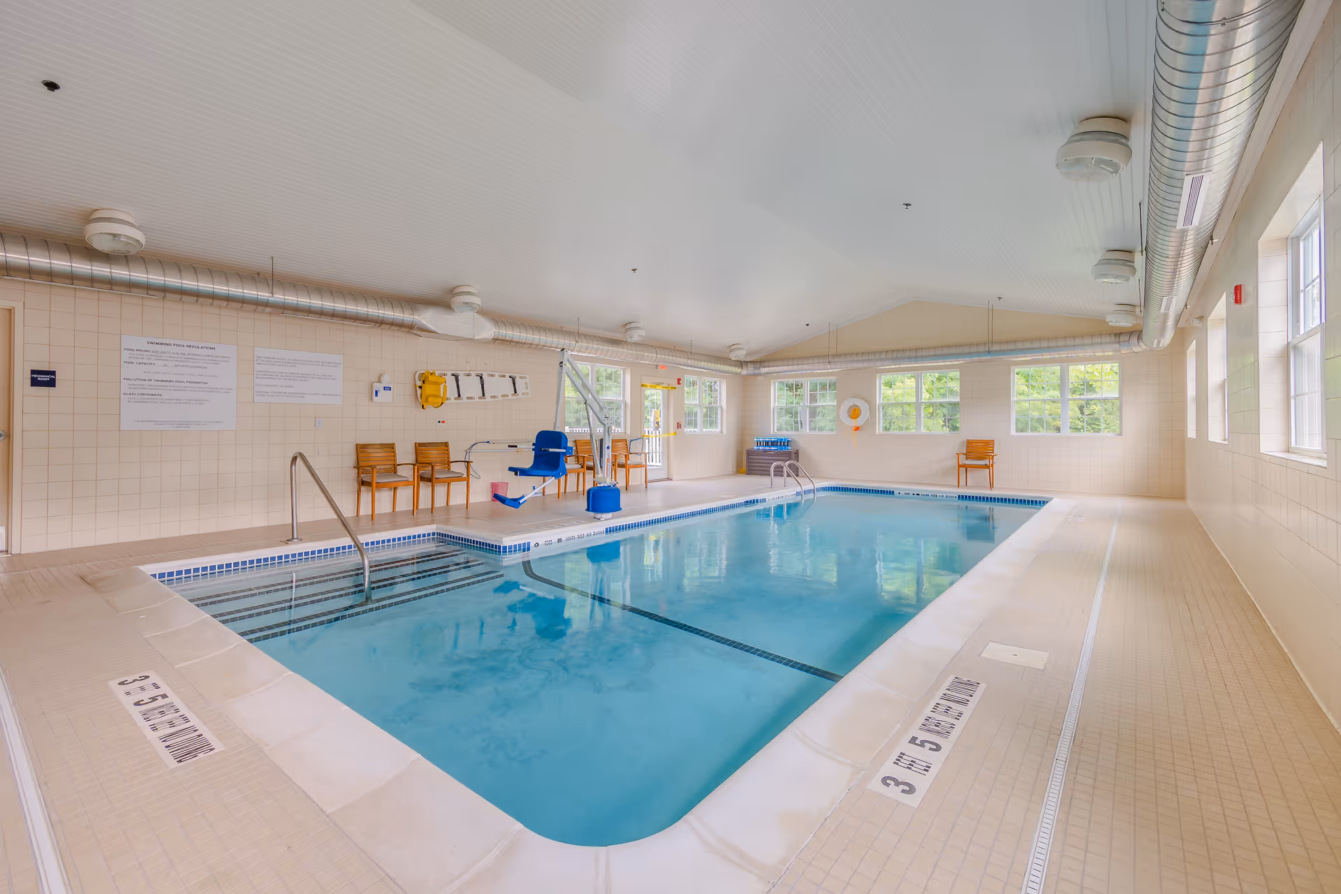 Bright indoor swimming pool with a handicap lift, chairs, and windows in a tiled room.
