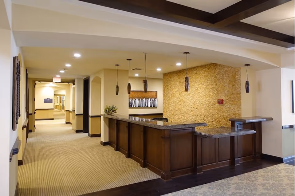 Interior view of a senior living facility reception area with a wooden front desk, pendant lights hanging from the ceiling, a textured stone accent wall behind the desk, and a hallway leading to other rooms.