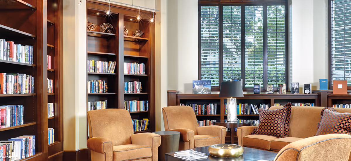 Cozy library room with wooden bookshelves filled with books, three upholstered armchairs, a sofa with patterned pillows, a round coffee table with a decorative bowl, and large windows with blinds letting in natural light.