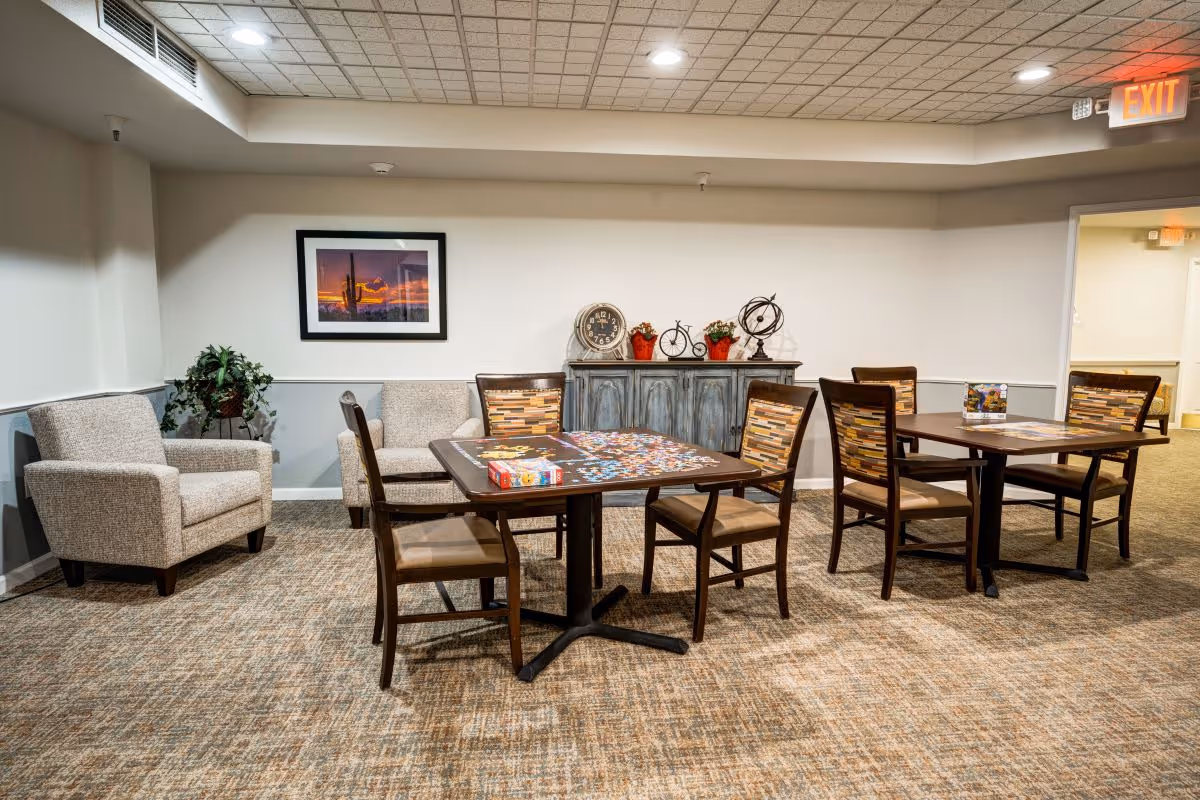 A well-lit common area with two tables and several chairs arranged around them. One table has a partially completed puzzle and a puzzle box on it. There are two upholstered armchairs in the corner next to a potted plant. A framed picture of a desert sunset hangs on the wall above a blue-gray sideboard decorated with a clock, small bicycle figurine, red flower pots, and a globe. An exit sign is visible above a doorway on the right.