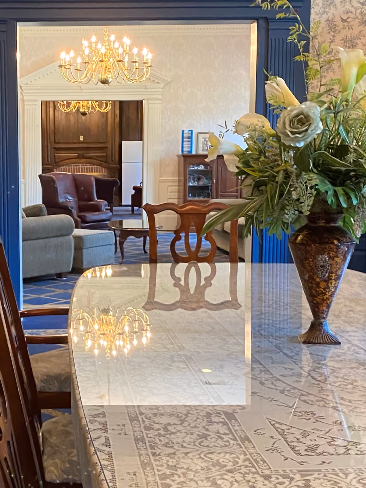 A polished dining table with a floral tablecloth and a large decorative vase filled with white flowers and greenery. In the background, there is a living room area with upholstered chairs, a wooden coffee table, and a chandelier hanging from the ceiling. The room features elegant wallpaper and wood paneling.