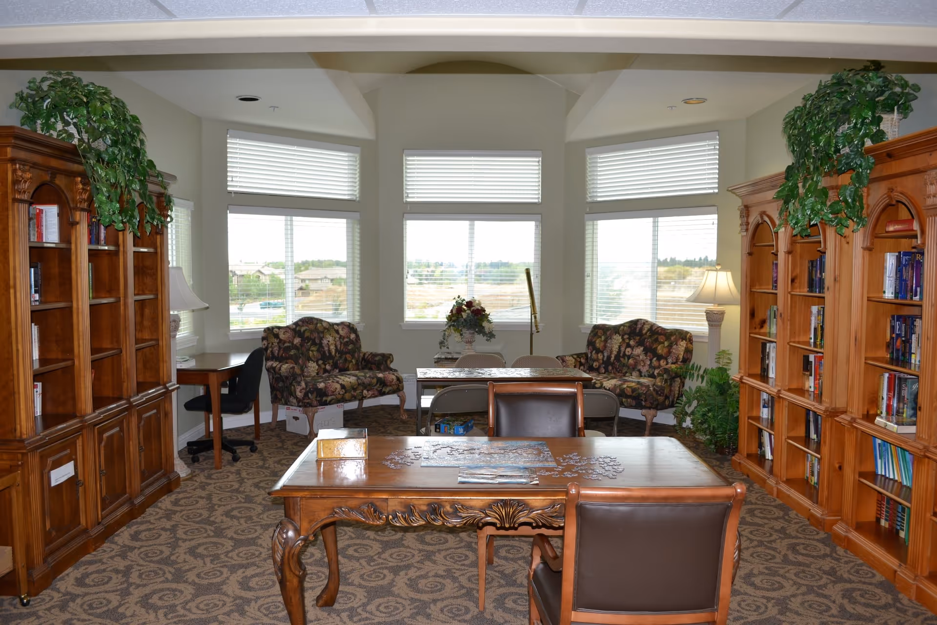 A bright communal library area with wooden bookshelves, plants, and cushioned seating near windows.