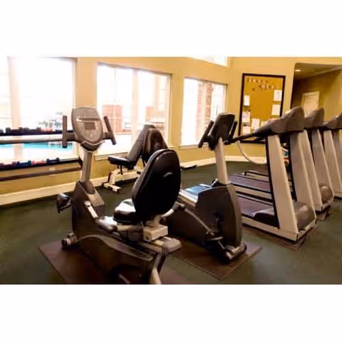 Indoor fitness room with exercise equipment including stationary bikes and treadmills, large windows letting in natural light, and a bulletin board on the wall.