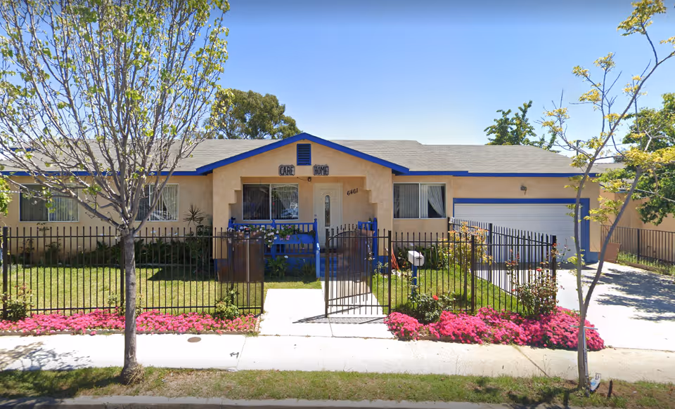 Single-story care home with beige exterior, blue trim, gated front yard, and pink flowers.