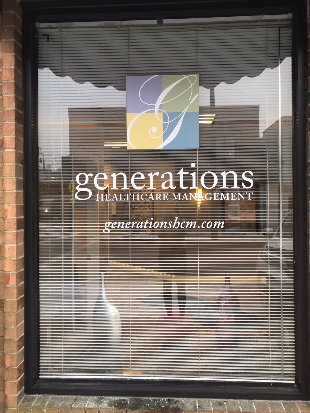 Window with closed blinds displaying the logo and name 'generations HEALTHCARE MANAGEMENT' along with the website 'generationshcm.com'. The logo features a stylized letter G in a square divided into four colored sections: purple, green, yellow, and blue. Reflections of the street and a person taking the photo are visible on the glass.