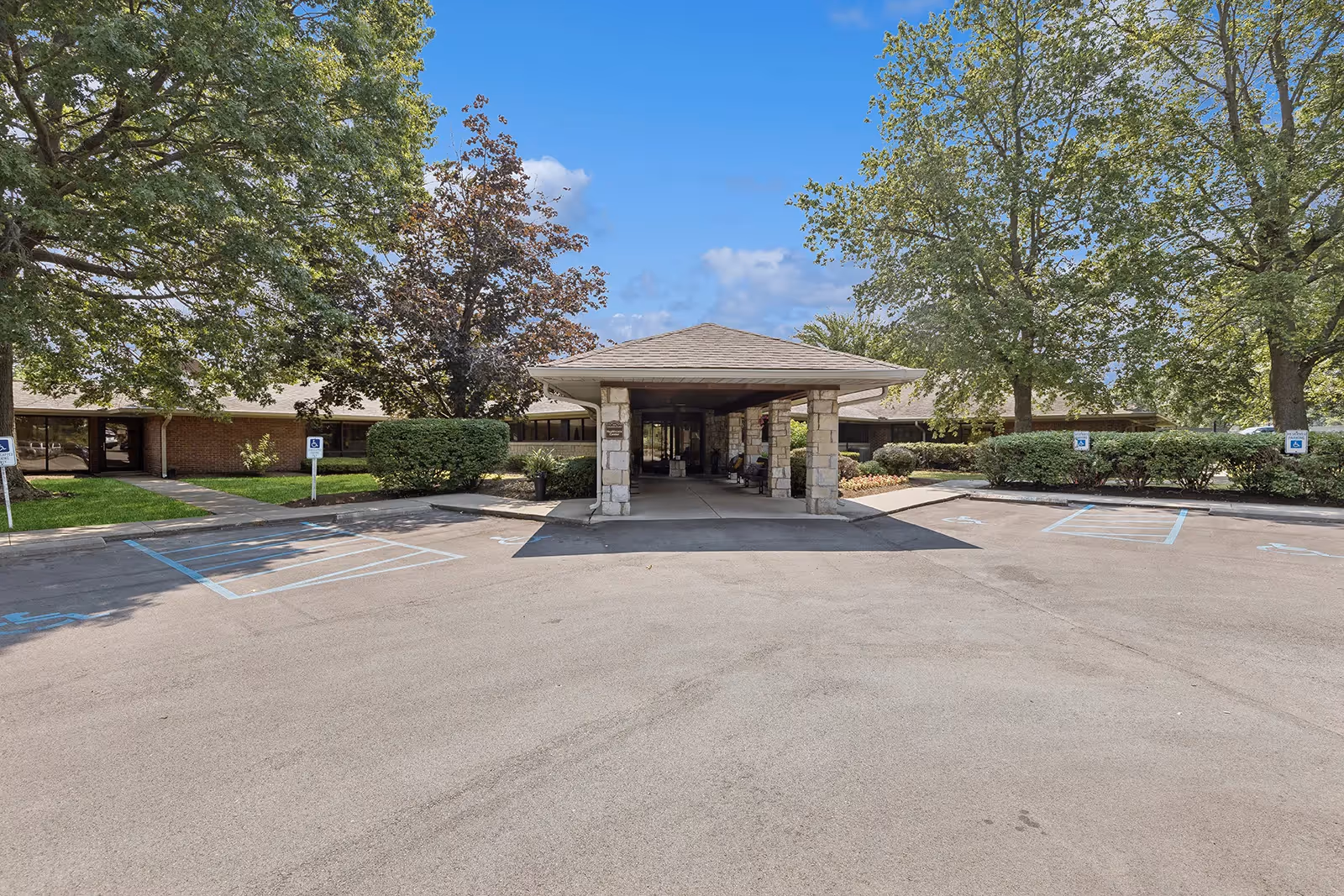 Covered entrance (porte-cochere) of a single-story senior living facility with handicap parking spaces and surrounding trees.