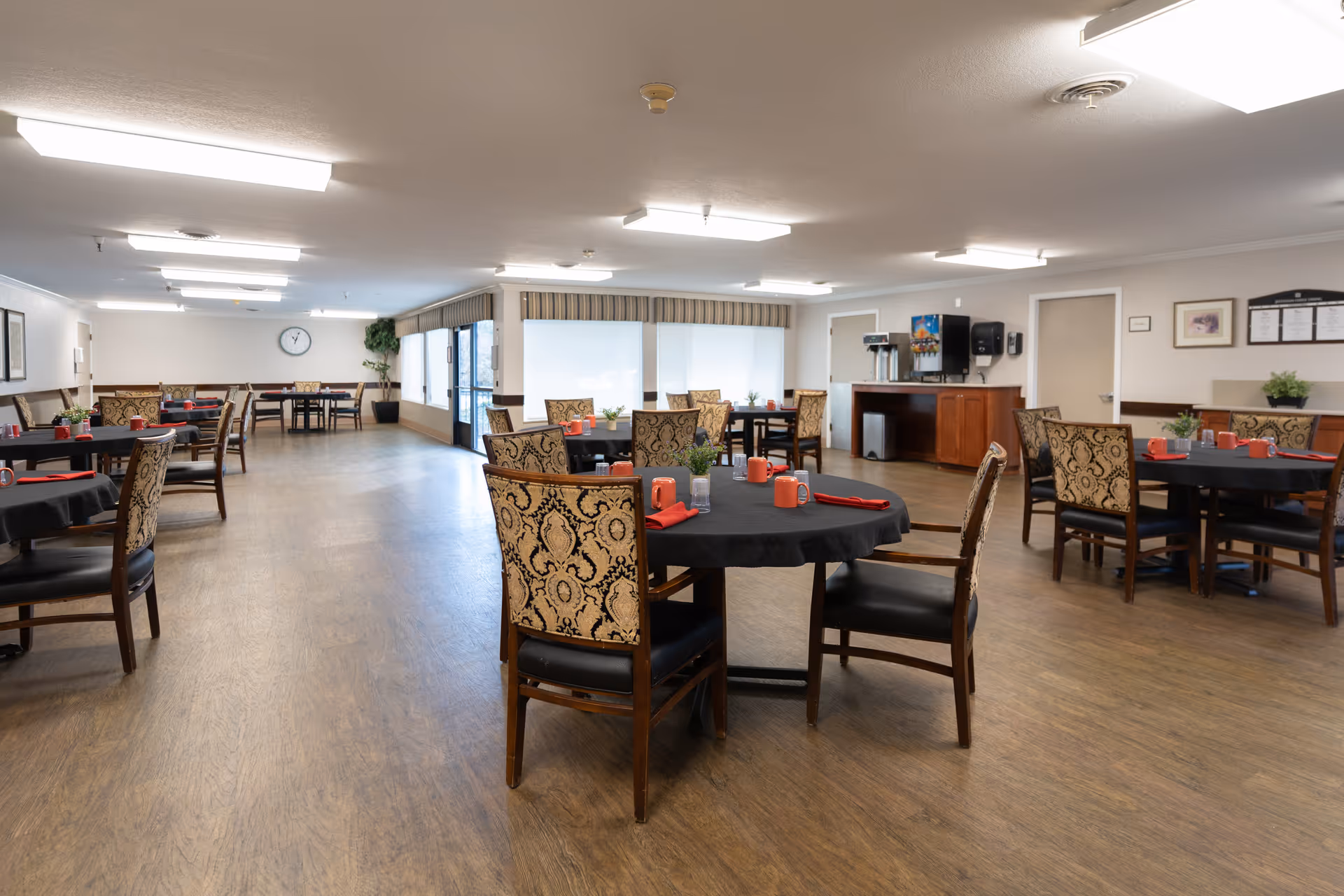 A spacious dining room in a senior living facility with multiple round tables covered with black tablecloths. Each table is set with red napkins, red mugs, and small potted plants. The room has wooden flooring, patterned chairs, large windows with blinds, and a beverage station with coffee dispensers against one wall.