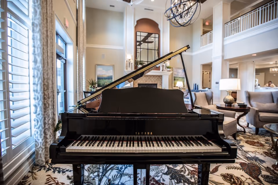 A black Yamaha grand piano in the center of a spacious, well-lit living room with high ceilings. The room features large windows with patterned curtains, comfortable armchairs, a patterned carpet, a fireplace with a mirror above it, and decorative lamps on side tables.