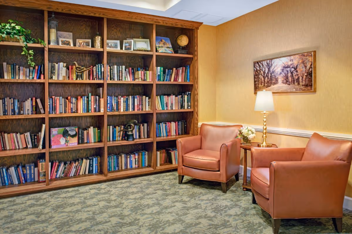 A cozy reading nook featuring a large wooden bookshelf filled with books and decorative items, two brown leather armchairs, a small wooden side table with a lamp and a flower arrangement, and a framed picture of a tree-lined path on the wall.