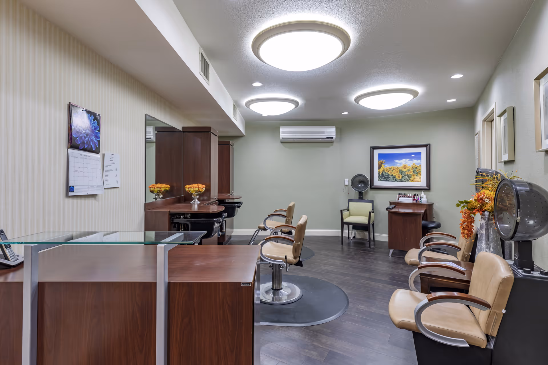 Interior view of a salon area with multiple styling chairs, mirrors, and hair drying stations. The room has modern circular ceiling lights, a wall-mounted air conditioner, a framed picture of sunflowers, and a small desk with nail polish and a fan. The floor is dark wood, and there are decorative flowers on the desk and near the hair dryers.