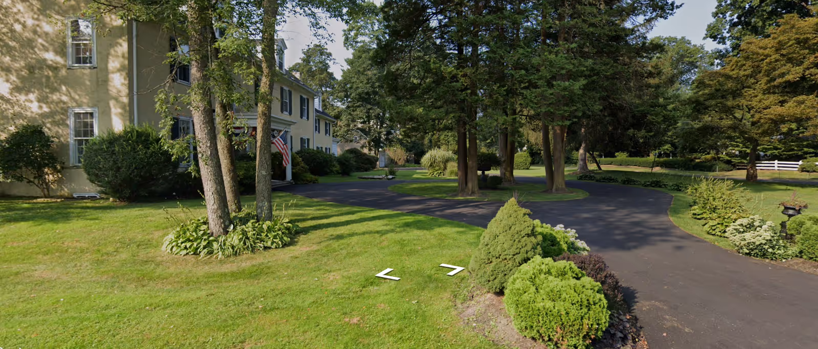Exterior view of a large yellow building with multiple windows and an American flag near the entrance, surrounded by green grass, trees, and shrubs along a curved paved driveway.