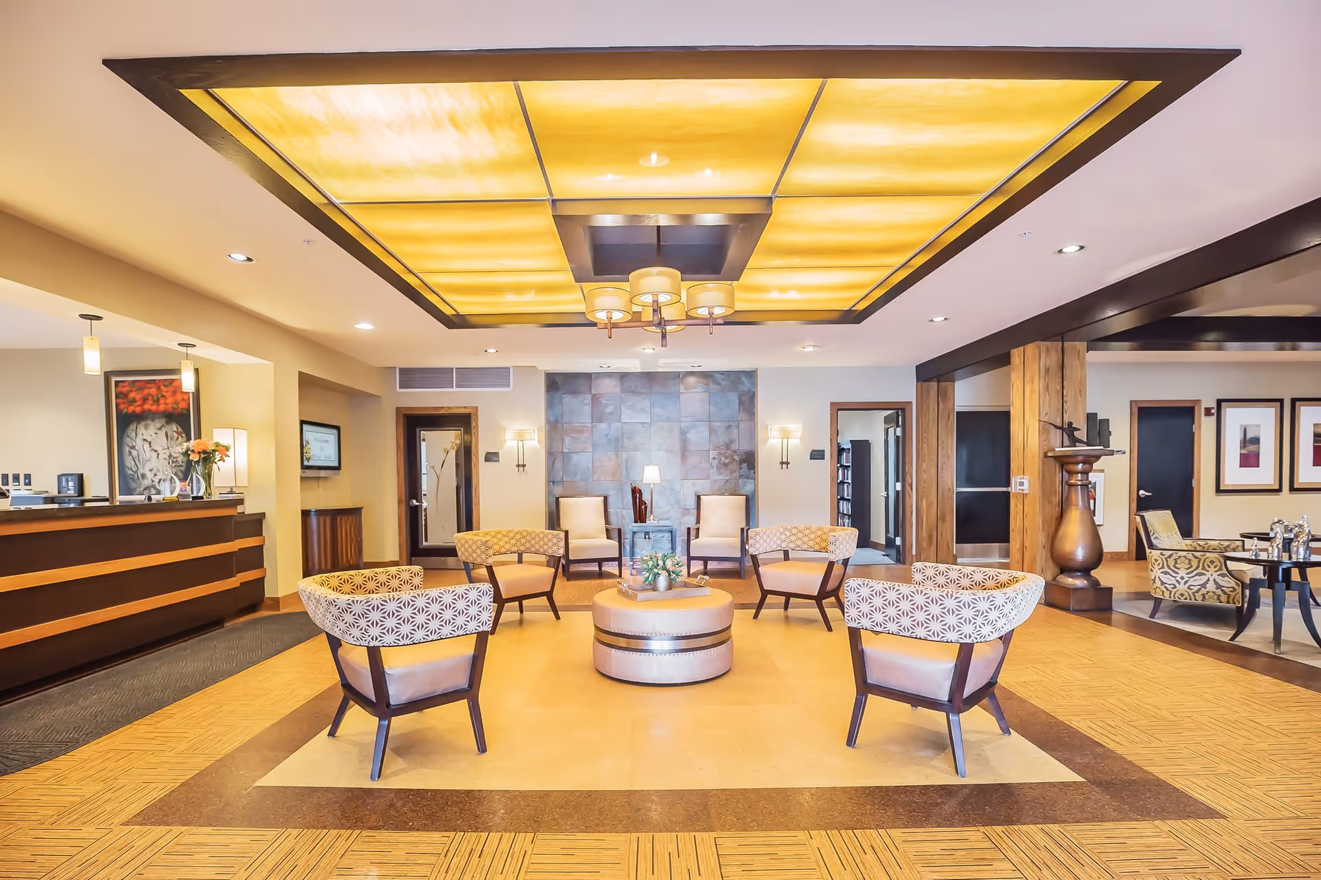 A spacious and well-lit senior living facility common area with a modern ceiling light fixture, several patterned armchairs arranged around a round ottoman with a small plant on it, a reception desk on the left, and additional seating and framed artwork in the background.