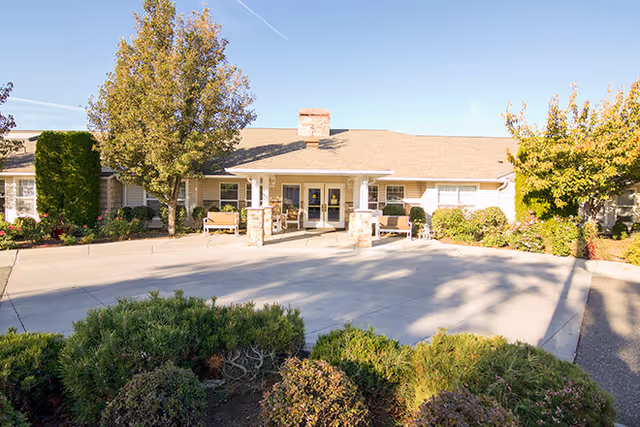 Single-story senior living facility entrance with a covered drop-off, benches, and landscaped shrubs under a clear sky.