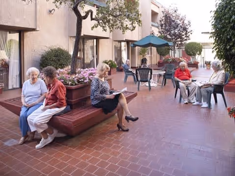 Several elderly people sitting and relaxing in an outdoor courtyard area of a senior living facility. The courtyard has brick flooring, potted plants, and patio furniture including chairs and tables with umbrellas. The building exterior is visible with windows and doors leading to the courtyard.