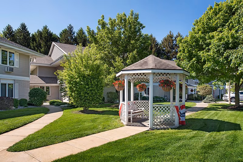 A well-maintained outdoor area at American House Spring Lake featuring a white gazebo decorated with hanging flower baskets and patriotic bunting. The gazebo is surrounded by lush green grass, trees, and paved walkways, with parts of the residential building visible in the background under a clear blue sky.