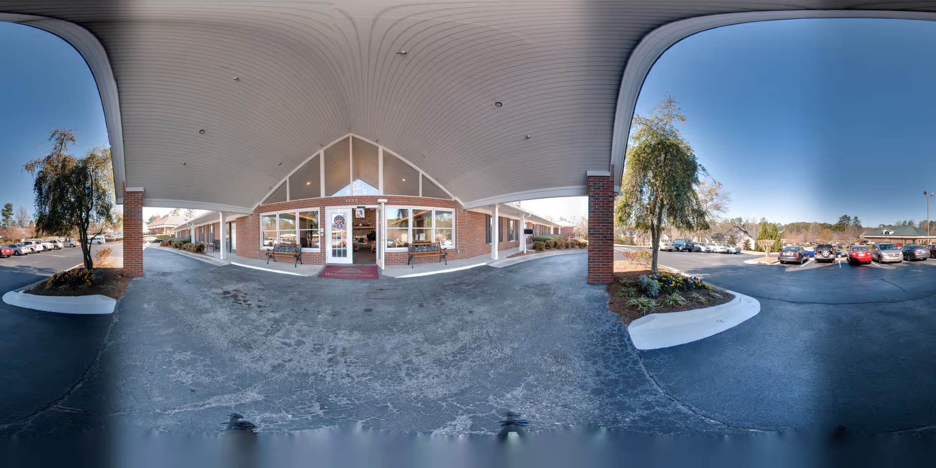 Covered entrance area of a senior living facility with a brick exterior, large windows, and a glass door. There are benches on either side of the entrance and landscaped areas with trees and flowers. A parking lot with several cars is visible on both sides under a clear blue sky.