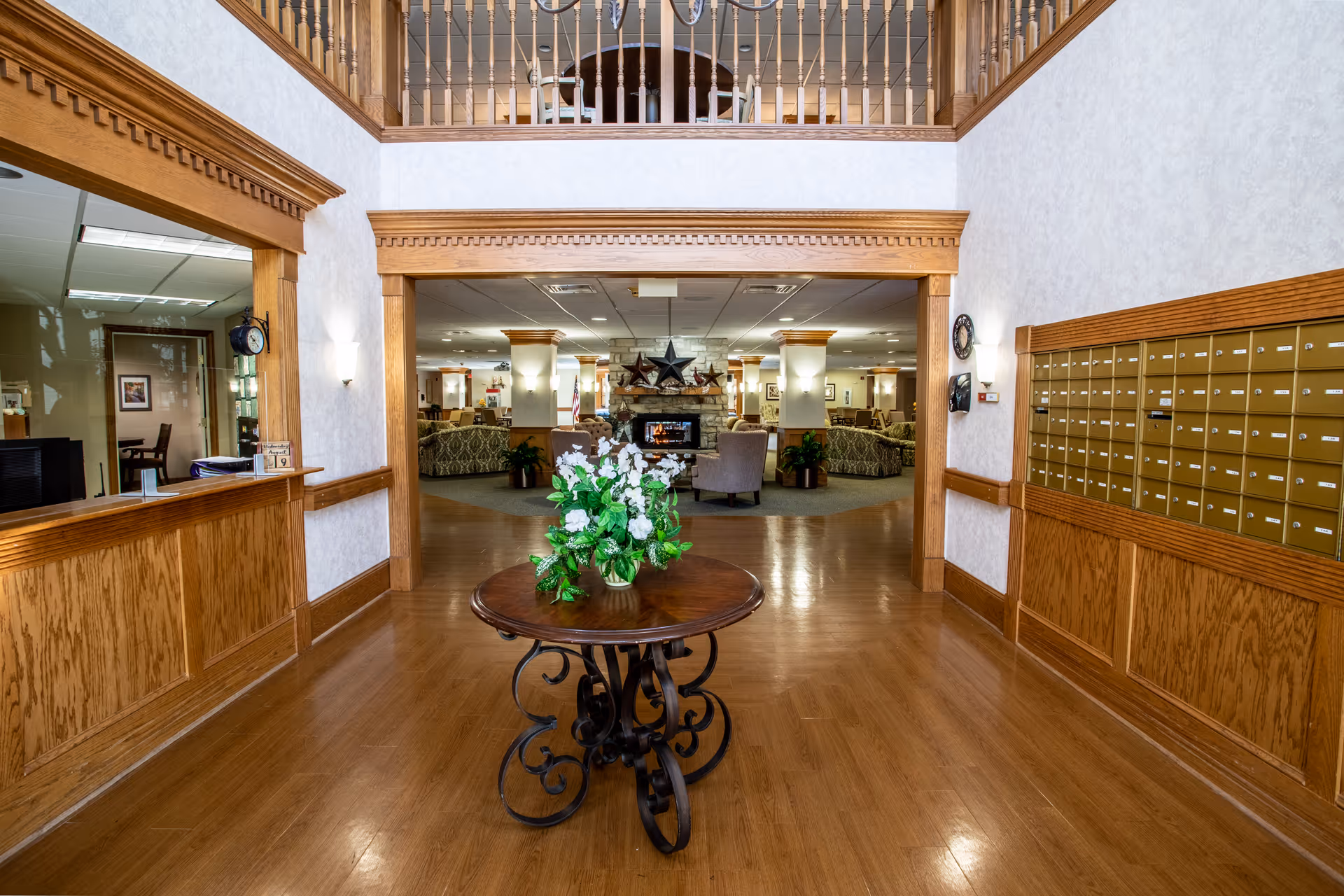Interior view of a senior living facility hallway with wooden floors and wood-paneled walls. A round wooden table with a floral arrangement is centered in the hallway. To the right, there is a wall of mailboxes. To the left, there is a reception window with a clock above it. The hallway opens into a spacious common area with comfortable seating and a stone fireplace decorated with stars.