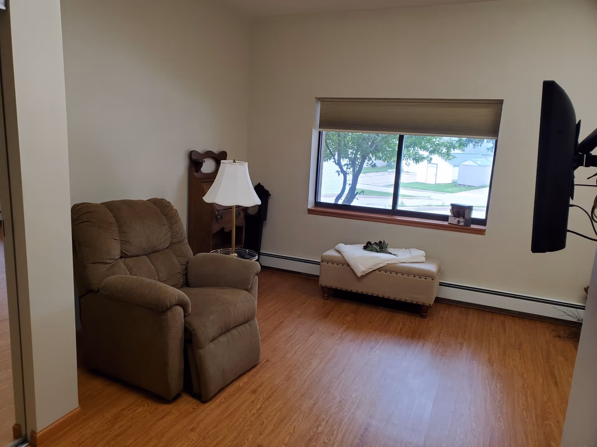 Small living room with a brown recliner, floor lamp, ottoman under a window, and a wall-mounted TV.