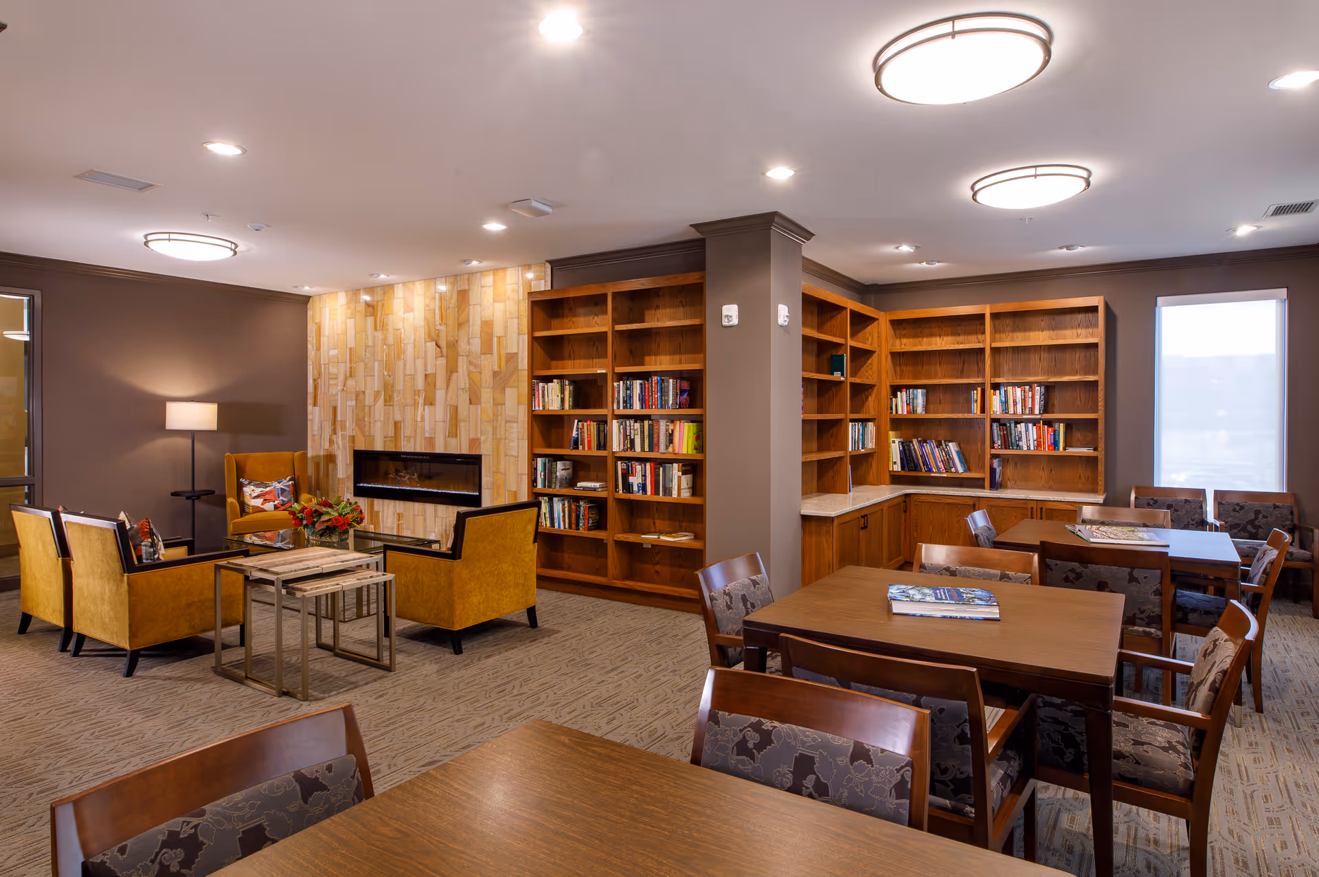A cozy common area in an assisted living facility featuring a seating area with mustard yellow armchairs around a glass coffee table in front of a modern fireplace with a tiled wall. Adjacent to the seating area are wooden bookshelves filled with books and several wooden tables with chairs, suitable for reading or social activities. The room is well-lit with ceiling lights and a floor lamp, and has large windows with blinds.
