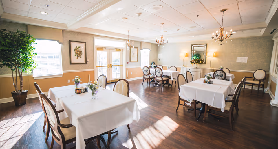 Bright dining room with wooden floors, several tables covered with white tablecloths, each set with small floral centerpieces. The room features large windows letting in natural light, decorative wall art, chandeliers, and a large mirror above a sideboard. There is a potted plant in the corner and double doors leading outside.