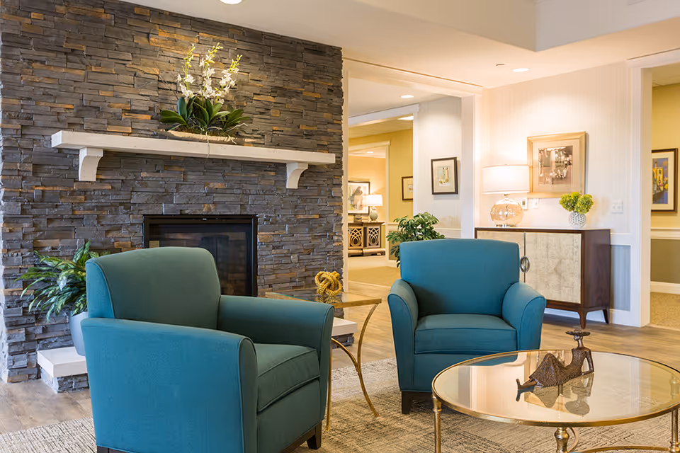 Cozy senior living lounge with two teal armchairs facing a stone fireplace and a glass-top coffee table.
