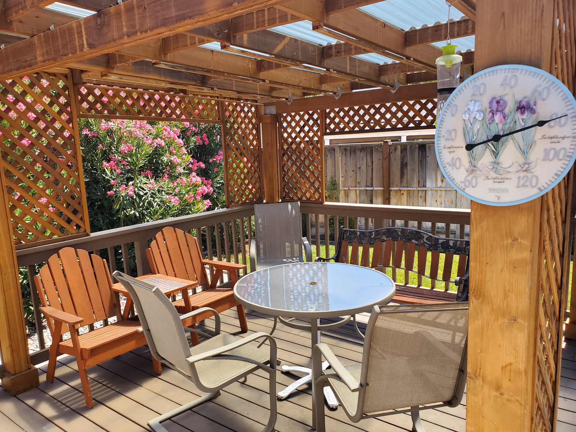 Outdoor covered patio area with wooden lattice walls and roof beams. There is a round glass table surrounded by four chairs, two wooden Adirondack chairs, and a wooden bench. Pink flowering bushes are visible outside the patio. A decorative thermometer with floral design is mounted on a wooden post.