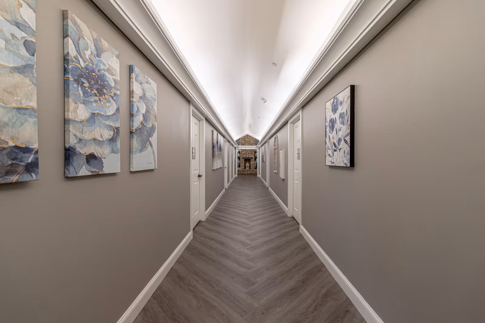 A long, well-lit hallway with gray walls and wood-patterned flooring. The ceiling has recessed lighting and crown molding. Both sides of the hallway feature white doors and framed floral artwork. At the end of the hallway, there is a stone fireplace.
