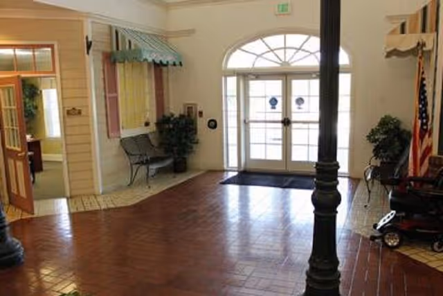 Interior view of an entrance lobby area with double glass doors letting in natural light. The floor is a combination of reddish-brown tiles and beige tiles. There are benches and potted plants along the walls, a black column in the foreground, and an American flag on the right side. A small awning is above a window on the left wall, and an open door leads to another room.