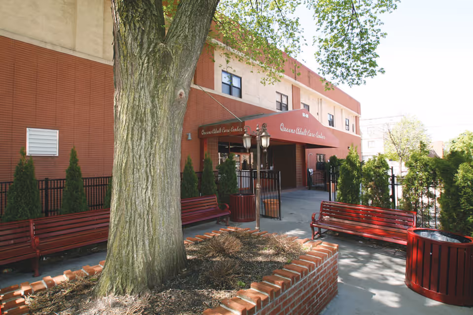 Outdoor seating area with red benches and a large tree in the center, located in front of the entrance to Queens Adult Care Center. The building has a red and beige exterior with a red awning displaying the facility name.