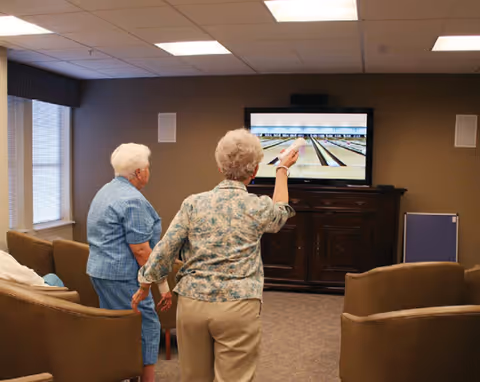 Two elderly women in a senior living common room playing a virtual bowling game shown on a television.