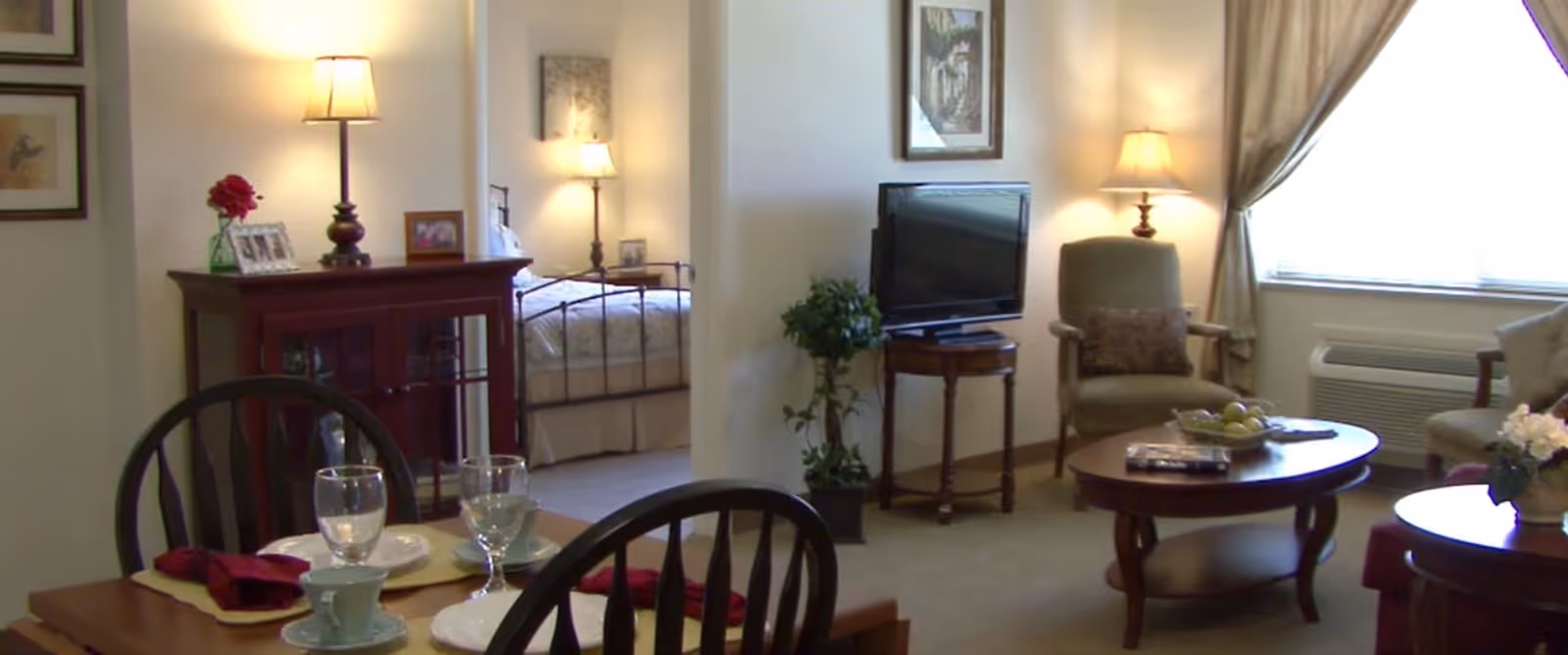 A cozy living and dining area in a senior living facility. The foreground shows a dining table set with plates, glasses, and cups. In the background, there is a living room with a TV on a small table, an armchair, a coffee table with a bowl of fruit, and a window with curtains. A bedroom with a bed and lamps is visible through an open doorway. The room is warmly lit with several lamps and decorated with framed pictures and plants.