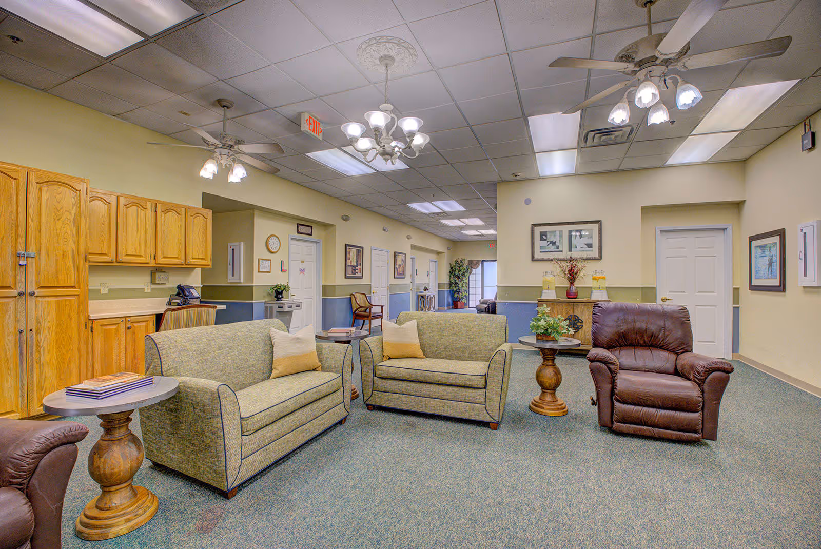 A well-lit common area in an assisted living facility featuring two green upholstered loveseats with beige pillows, a brown leather recliner, wooden side tables with plants and books, wooden cabinets along one wall, and framed artwork on the walls. The ceiling has multiple light fixtures including chandeliers and ceiling fans. Doors and hallway are visible in the background.