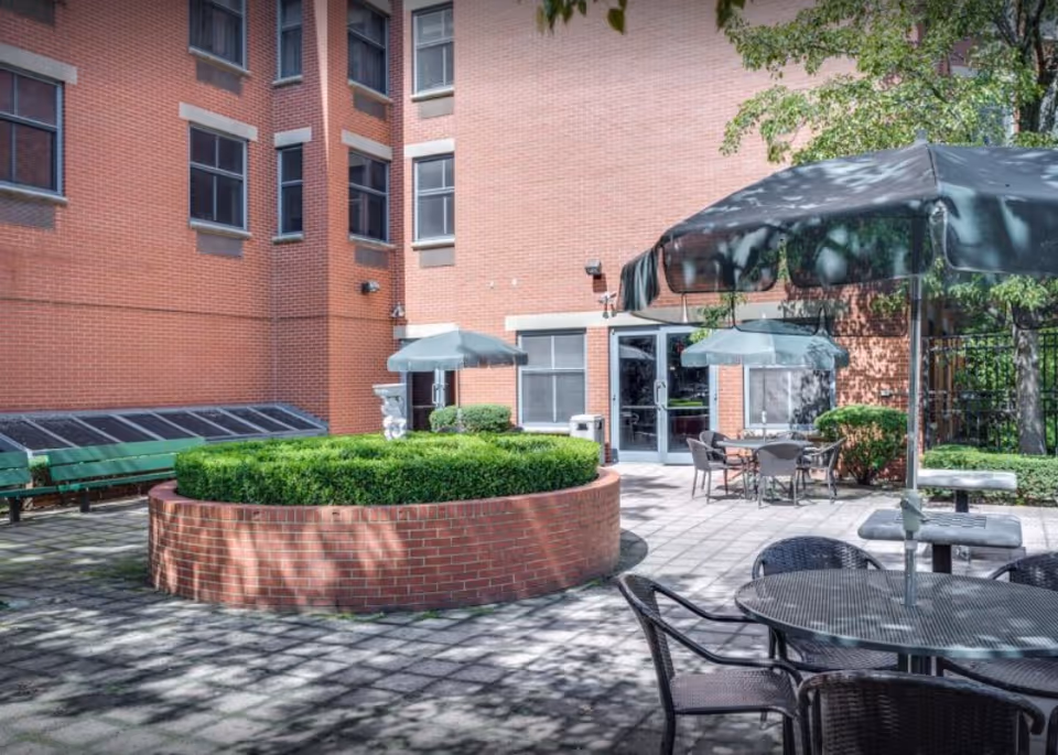 Outdoor courtyard area with round brick planter filled with green bushes, several tables with umbrellas, chairs, and a brick building with multiple windows in the background.