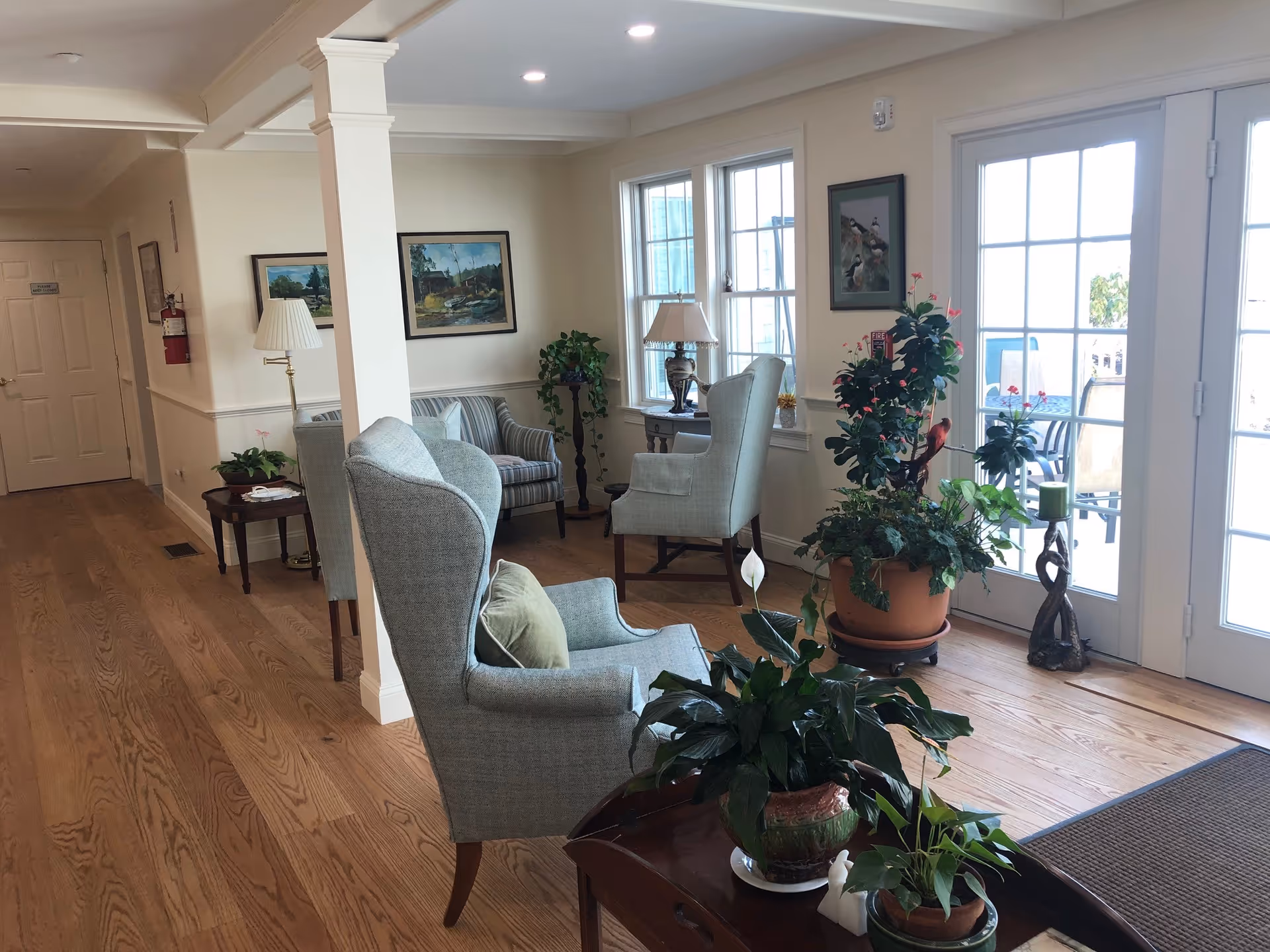 A bright and cozy sitting area in a senior living facility with wooden floors, light-colored walls, and large windows letting in natural light. The room features several upholstered armchairs and a striped loveseat, along with multiple potted plants and framed artwork on the walls. A glass door leads to an outdoor patio area with chairs and a table.