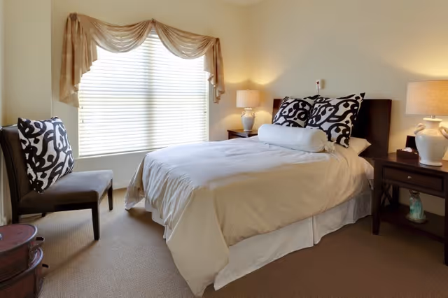 A neatly made bed with white bedding and two decorative black and white pillows in a bedroom. There are two bedside tables with matching lamps on either side of the bed. A chair with a black and white patterned cushion is positioned near a window with blinds and a beige valance. The room has beige walls and carpeted floor.