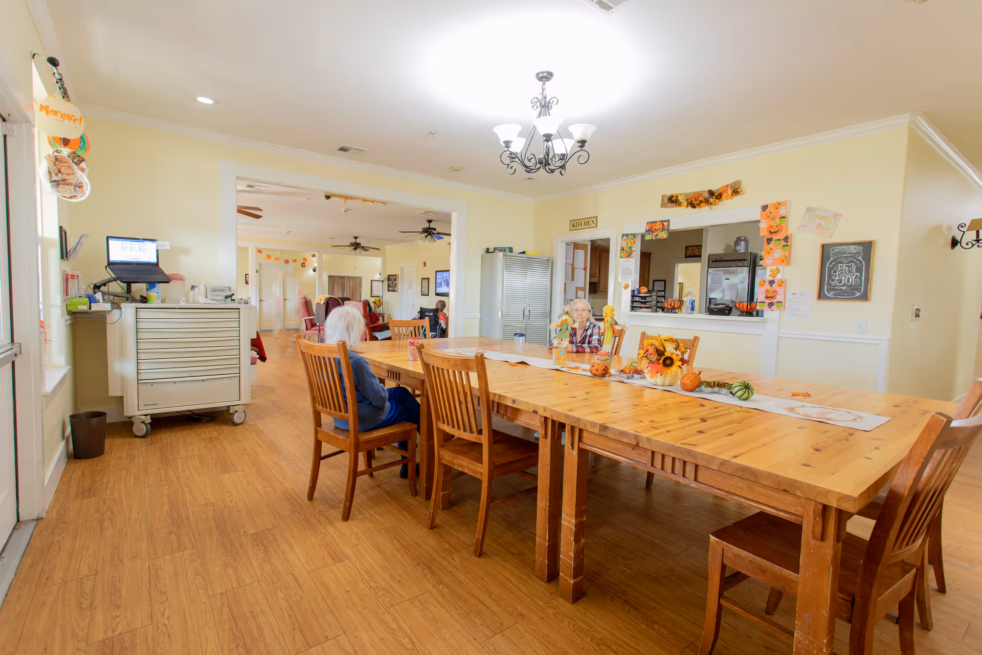 A bright dining area in Mercy House Temple with wooden tables and chairs. Two elderly women are seated at the table, which is decorated with autumn-themed items including small pumpkins and flowers. The room has light yellow walls, a chandelier, and a serving window labeled 'Kitchen' with festive decorations around it. In the background, there is another room with more seating and ceiling fans.