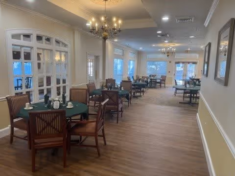 A spacious dining room with multiple round tables covered with green tablecloths and set with cups and napkins. Wooden chairs surround the tables. The room features chandeliers hanging from the ceiling, large windows letting in natural light, and light-colored walls with decorative molding.