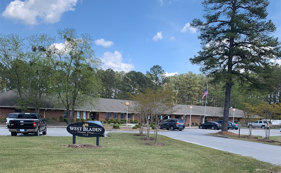 Exterior view of West Bladen Assisted Living and Memory Care facility showing a single-story brick building with a parking lot in front. Several cars are parked, and there are trees and a grassy area with a sign that reads 'West Bladen Assisted Living 714'. The sky is blue with some clouds.