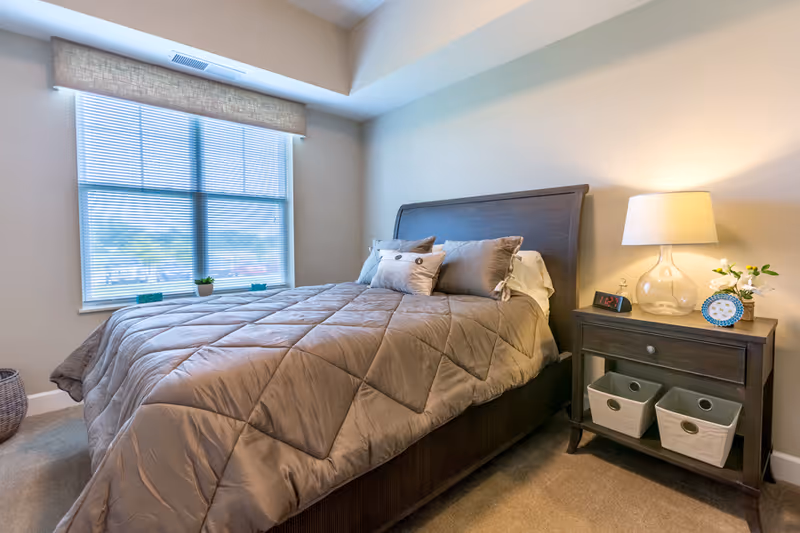 A cozy bedroom with a large window covered by blinds and a beige valance. The bed has a dark wooden frame and is made with a taupe quilted comforter and several pillows. Next to the bed is a dark wooden nightstand with a glass lamp, a digital alarm clock, a small plant, and a framed photo. The room has beige walls and carpeted floor.