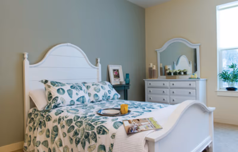 A bright bedroom with a white wooden bed frame and matching dresser with mirror. The bed is made with a white and green leaf-patterned quilt and pillows. A tray with a yellow cup and a magazine rests on the bed. There is a small side table with a framed photo and a blue vase. A window with white blinds lets in natural light, and a green plant sits on the windowsill.