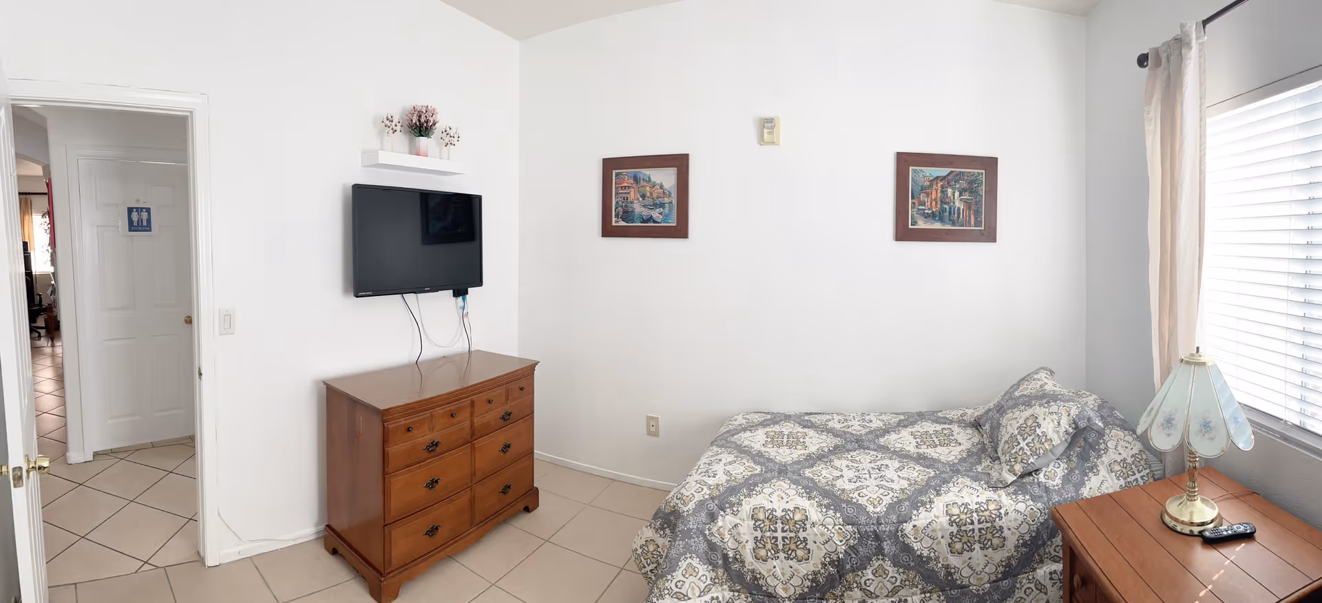 A small bedroom with a single bed covered in a patterned gray and white bedspread. Next to the bed is a wooden nightstand with a decorative lamp and a remote control. On the opposite wall, there is a wooden dresser with a flat-screen TV mounted above it and a small shelf with decorative items. Two framed pictures hang on the wall above the bed. The room has tiled floors and a window with white blinds and beige curtains. An open door leads to a hallway with tiled floors and a restroom sign visible on a door in the background.