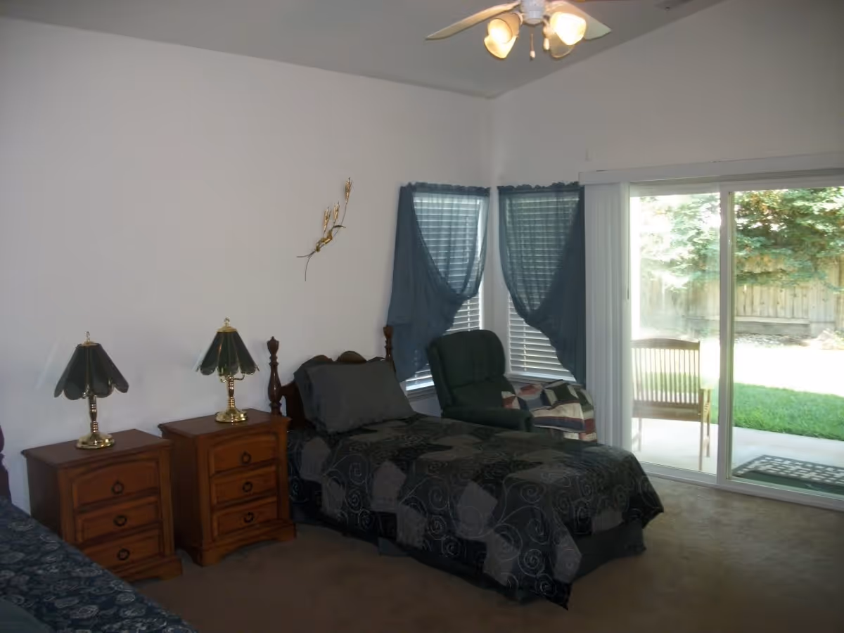 A bedroom with a single bed covered in a dark patterned bedspread, two wooden nightstands with matching lamps, a green armchair with a quilt draped over it, two windows with blue curtains, and a sliding glass door leading to a small outdoor patio with a wooden chair and green grass.