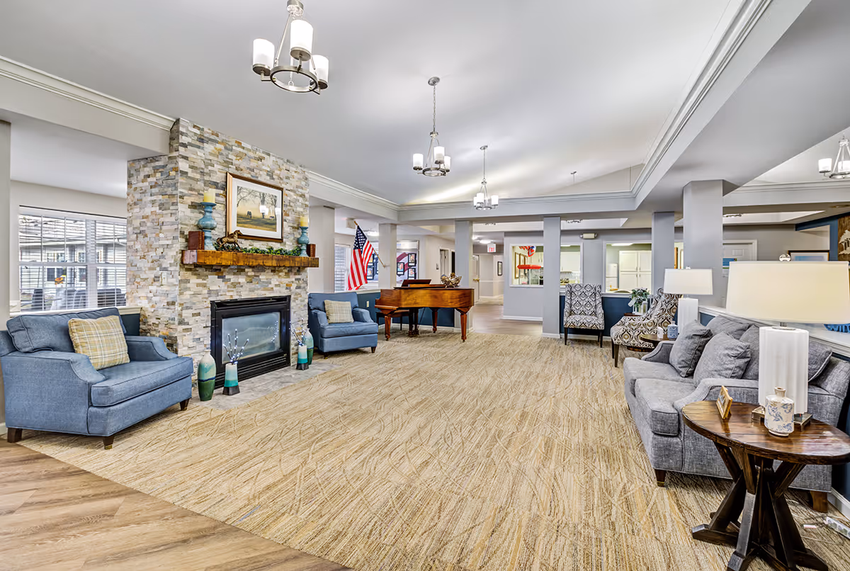 A spacious and well-lit living room area in a senior living facility featuring a stone fireplace with a wooden mantle, two blue armchairs with plaid pillows, a gray sofa with multiple cushions, two patterned chairs, a wooden side table with a lamp and decorative items, and a grand piano in the background. The room has beige carpet flooring, multiple ceiling light fixtures, and an American flag near the piano.