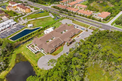 Aerial view of a senior living facility surrounded by greenery, roads, and nearby residential buildings. The facility has a large building with a brown roof, parking areas, and landscaped grounds including a pond and trees.