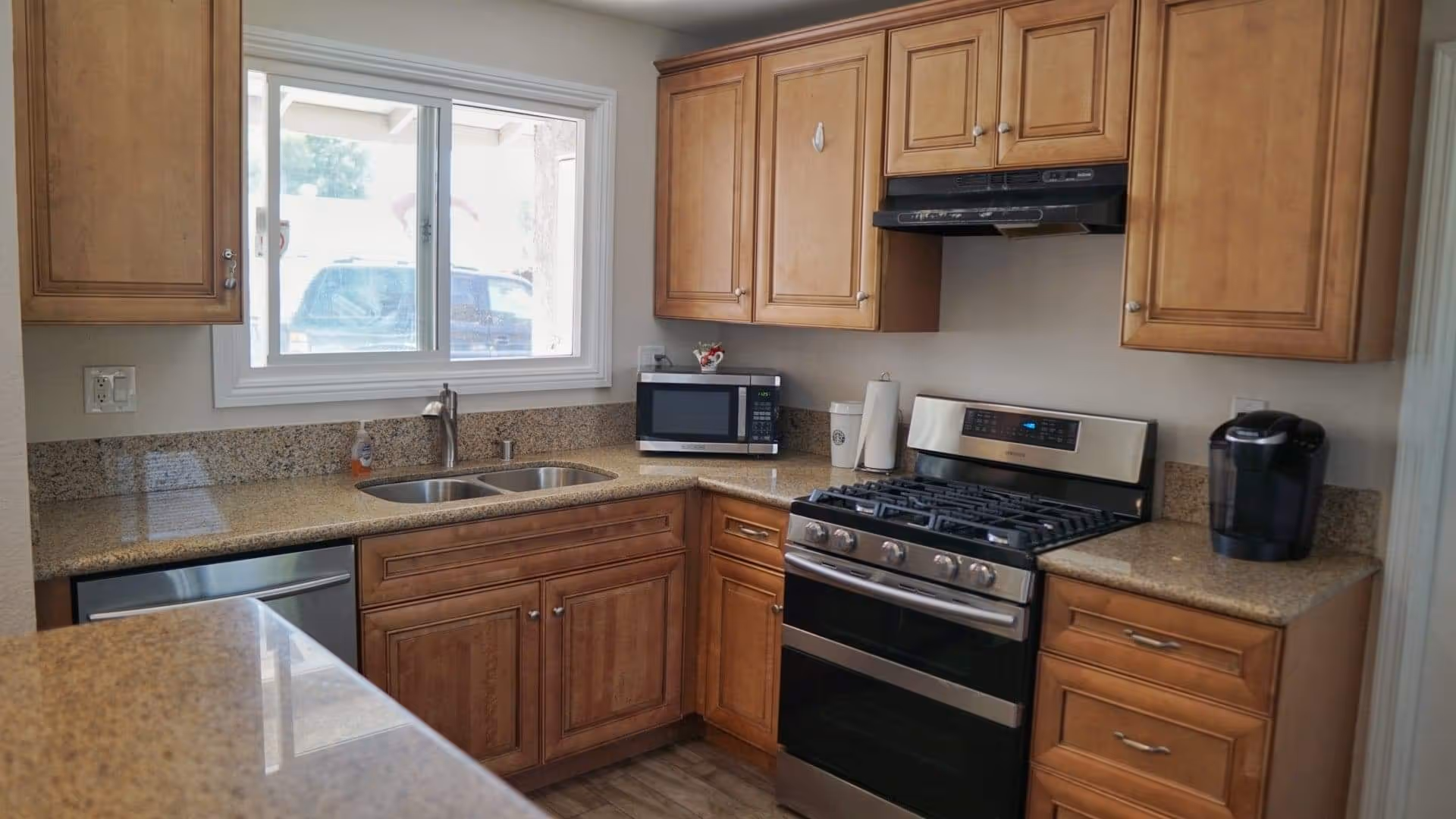 A kitchen with wooden cabinets, granite countertops, a double sink under a window, a microwave, a gas stove with an oven, a dishwasher, and a coffee maker on the counter.