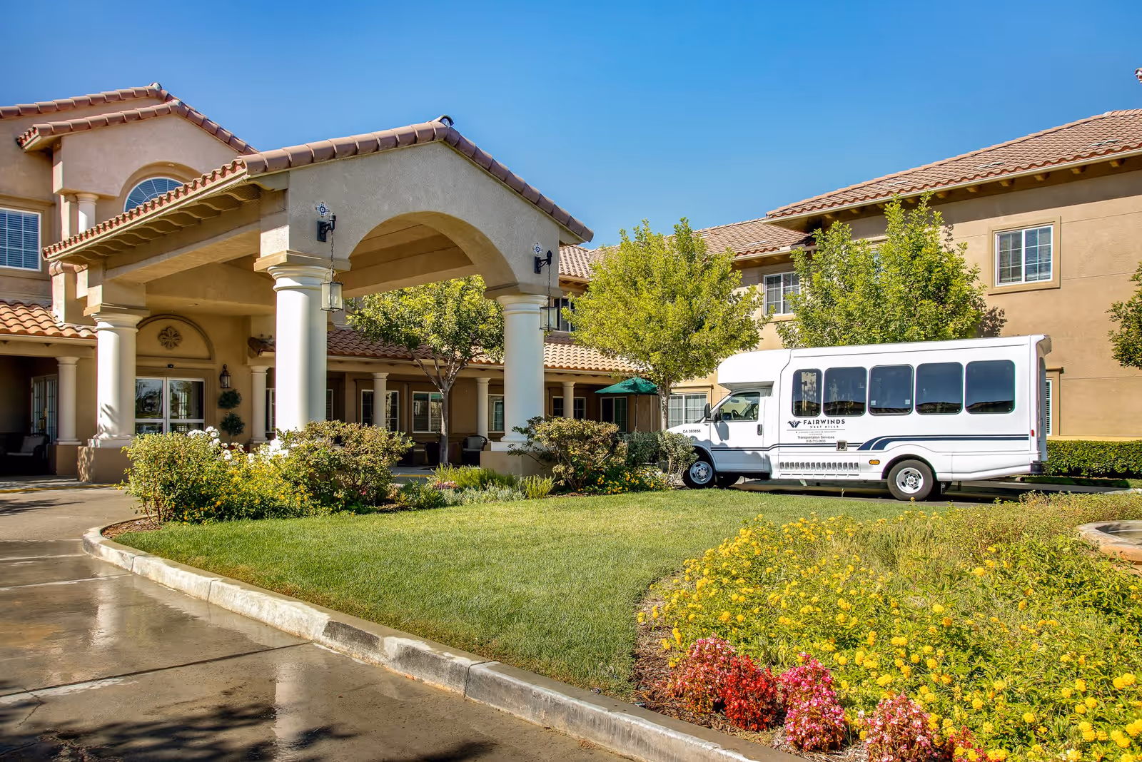 Exterior view of Fairwinds - West Hills senior living facility showing the entrance with a covered driveway supported by columns, a white shuttle bus parked nearby, green lawn, and colorful flower beds under a clear blue sky.