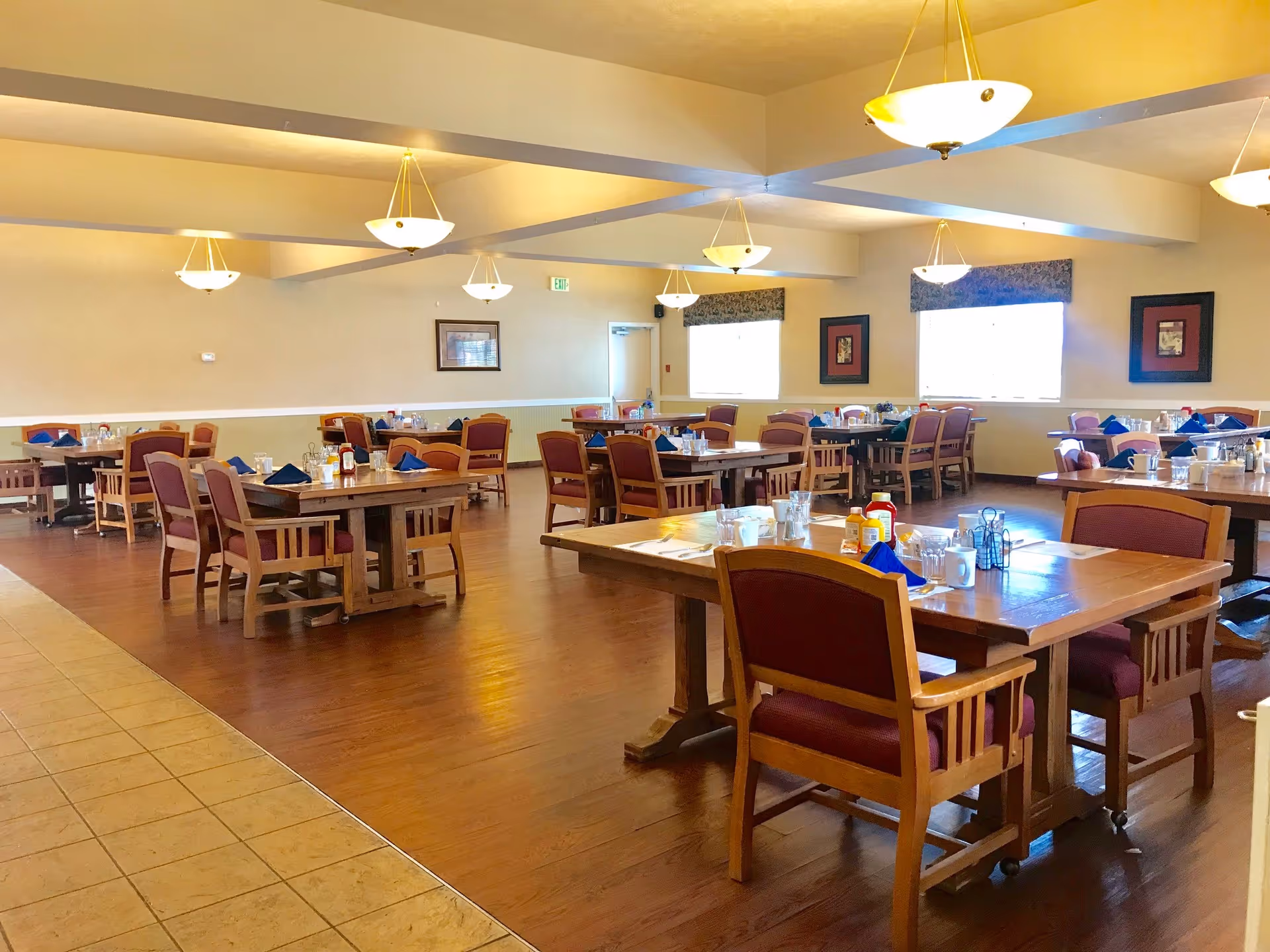 A spacious dining room in an assisted living facility with multiple wooden tables and chairs arranged neatly. Each table is set with glasses, mugs, napkins, and condiments. The room has warm lighting from ceiling fixtures and windows with valances, and framed artwork on the walls.