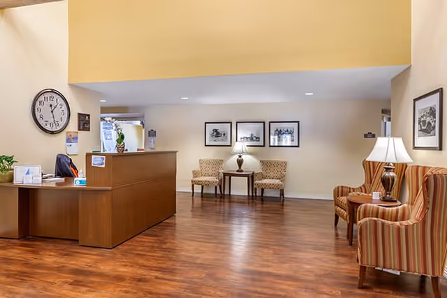 Reception area with a wooden desk, a clock on the wall, two beige patterned chairs with a small table and lamp between them, and two striped armchairs with a lamp on a side table in a room with wooden flooring and framed pictures on the walls.