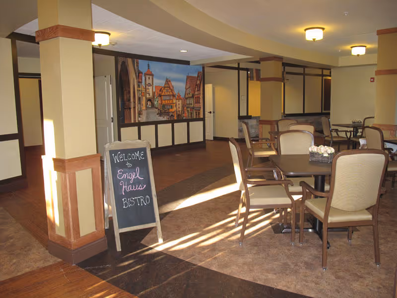 Interior view of a senior living facility dining area with several tables and chairs arranged neatly. A chalkboard sign near a pillar reads 'Welcome to Engel Haus Bistro.' The walls are decorated with a large mural depicting a quaint European village scene. The room is well-lit with ceiling lights and natural light coming from the left side.